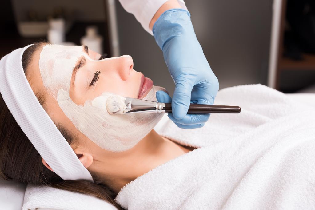 Kosmetikerin trägt Kosmetikmaske auf Gesicht einer Frau im Schönheitssalon auf - Foto, Bild