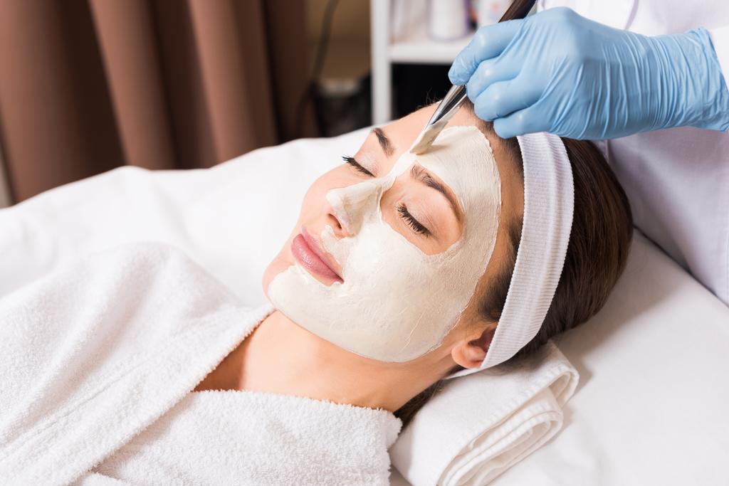Kosmetikerin trägt Kosmetikmaske mit Kosmetikpinsel auf Frau auf halbem Gesicht im Schönheitssalon auf - Foto, Bild