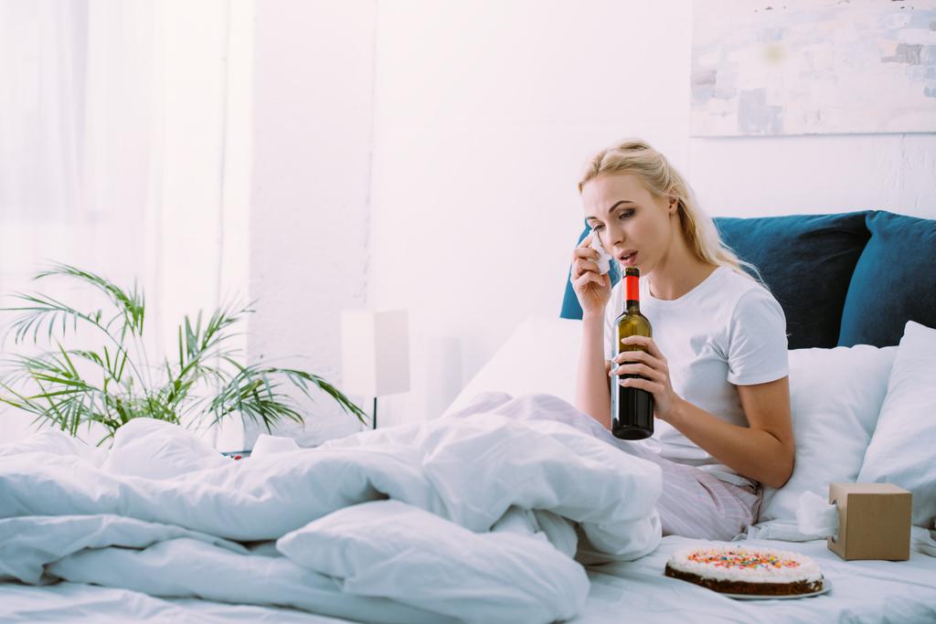 Traurige Frau mit Flasche Wein wischt sich Tränen, während sie Geburtstag allein im Bett feiert - Foto, Bild