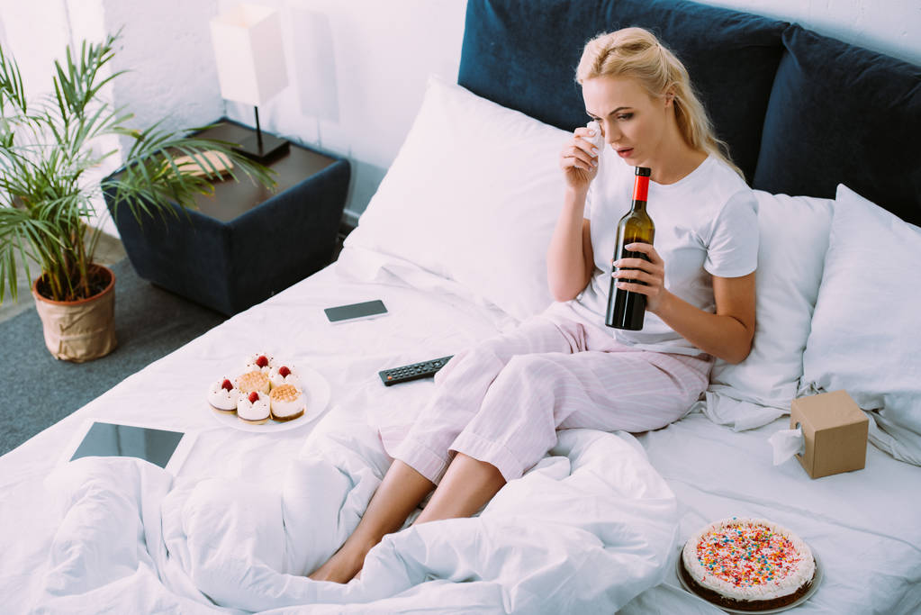 一人ぼっちでベッドで誕生日を祝っている間涙を拭くワインのボトルを持つ女性を混乱させる - 写真・画像