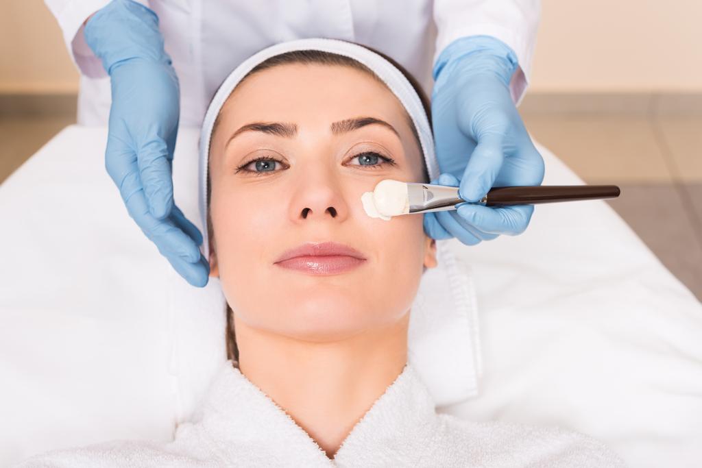 esteticista aplicando máscara cosmética com escova cosmética no rosto da mulher no salão de beleza
 - Foto, Imagem