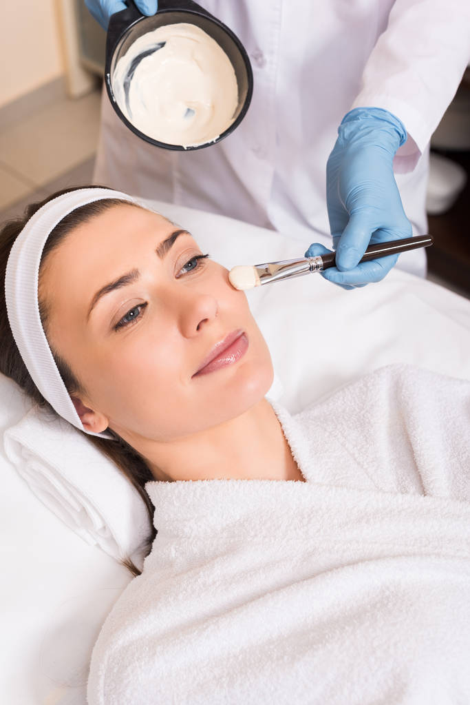 esteticista aplicando máscara cosmética na bochecha da mulher com escova cosmética no salão de beleza
 - Foto, Imagem