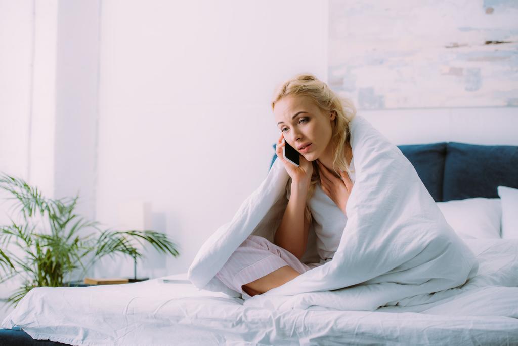 αναστατωμένος γυναίκα που καλύπτονται μέσα κουβέρτα μιλώντας στο smartphone στο κρεβάτι στο σπίτι με αντίγραφο χώρου - Φωτογραφία, εικόνα