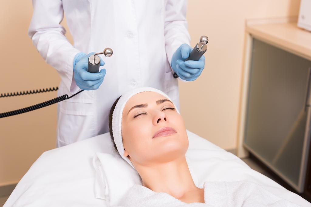 esteticista segurando massageador sobre mulher rosto no salão de beleza
 - Foto, Imagem