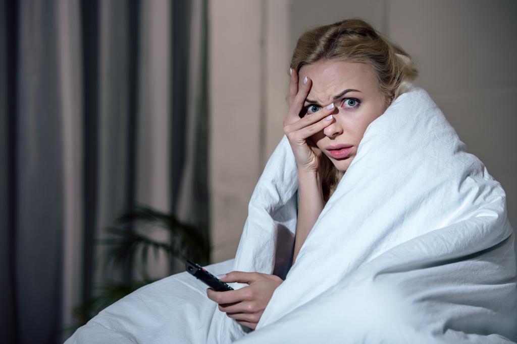 vyděšená žena pokrývající obličej s rukou při sledování televize v posteli doma - Fotografie, Obrázek