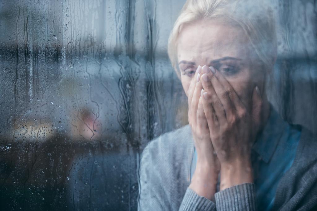 smutny Dorosła kobieta płacze i obejmujących twarz rękami w domu przez okno z deszczu - Zdjęcie, obraz
