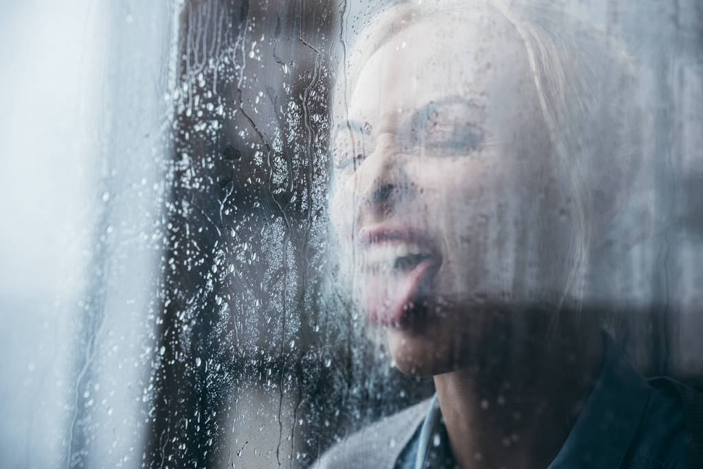 seçici odak kızgın yetişkin kadın dilini dışarı ve bağrışmalar evde yağmur damlaları ile pencereden yapışmasını. - Fotoğraf, Görsel