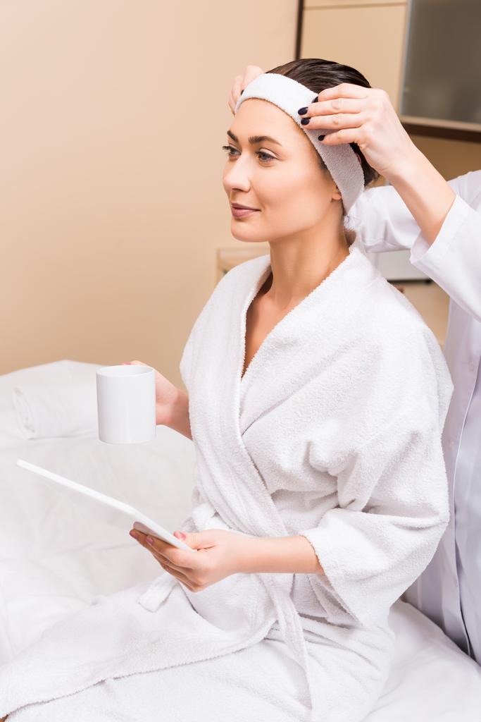 Nainen istuu, tilalla kuppi ja käyttää digitaalista tablettia, kun taas kosmetologi korjaa hänen hiusnauha kauneushoitola
 - Valokuva, kuva