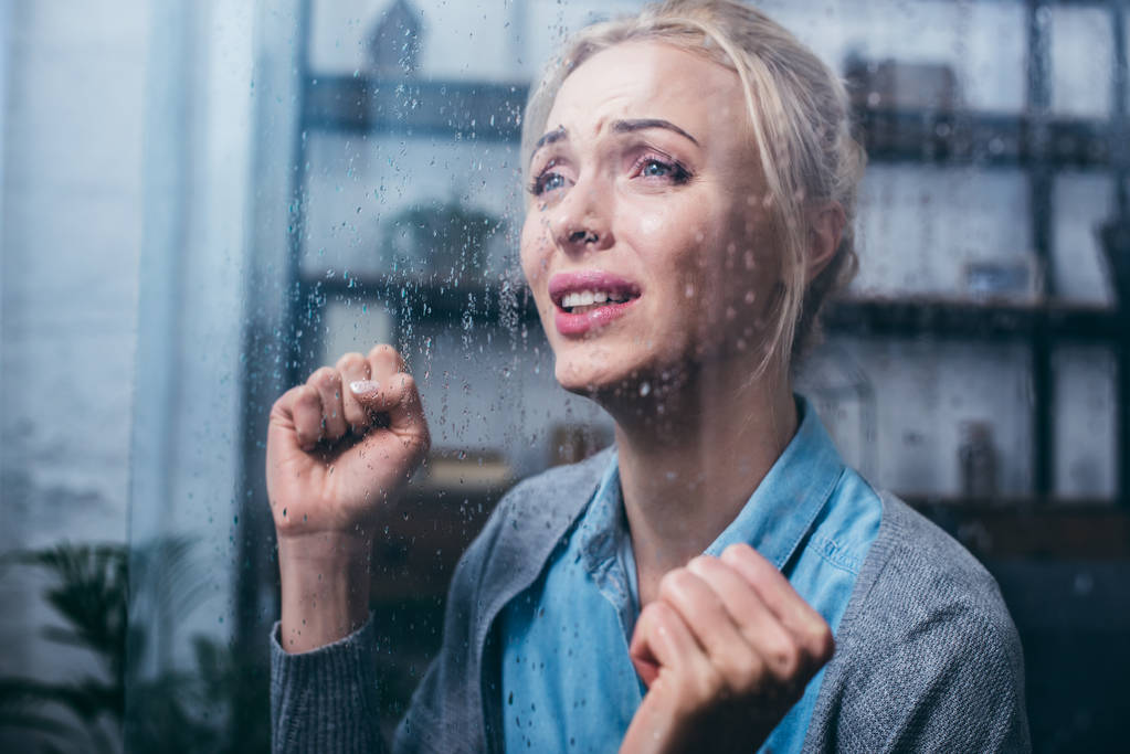 smutny Dorosła kobieta płacze z zaciśniętymi pięściami w domu przez okno z deszczu - Zdjęcie, obraz