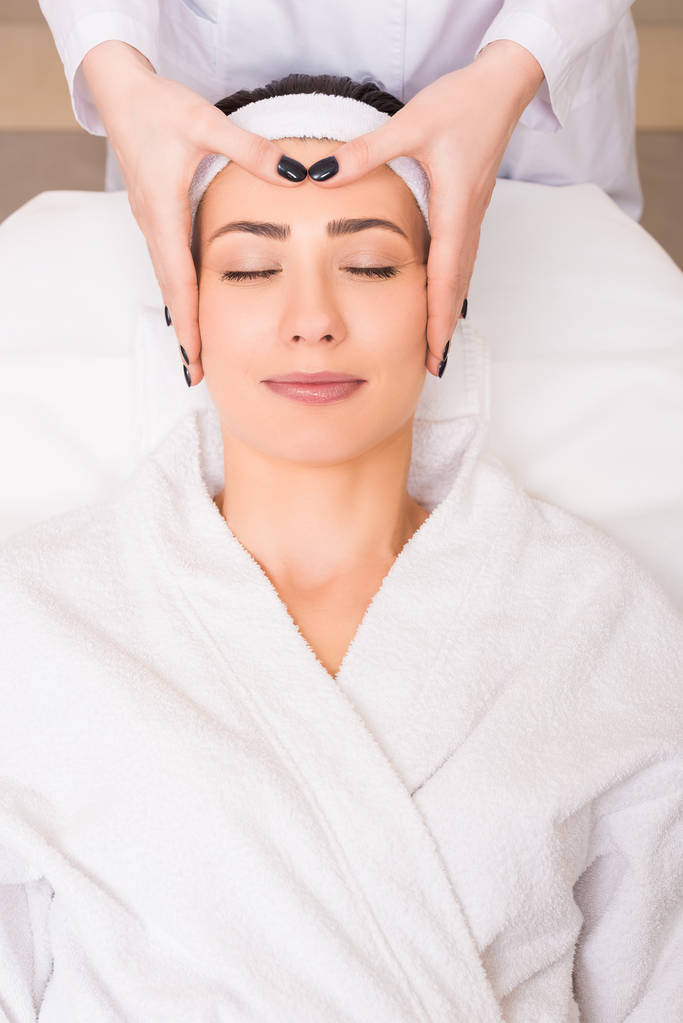 schoonheidsspecialist doen handmatige gezicht massage voor vrouw liggen op badjas en hairband op de salon van de schoonheid - Foto, afbeelding