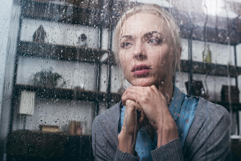 θλιβερή ενήλικη γυναίκα με σταυρωμένα χέρια να κοιτάμε μακριά στο σπίτι μέσα από το παράθυρο με τις σταγόνες της βροχής - Φωτογραφία, εικόνα