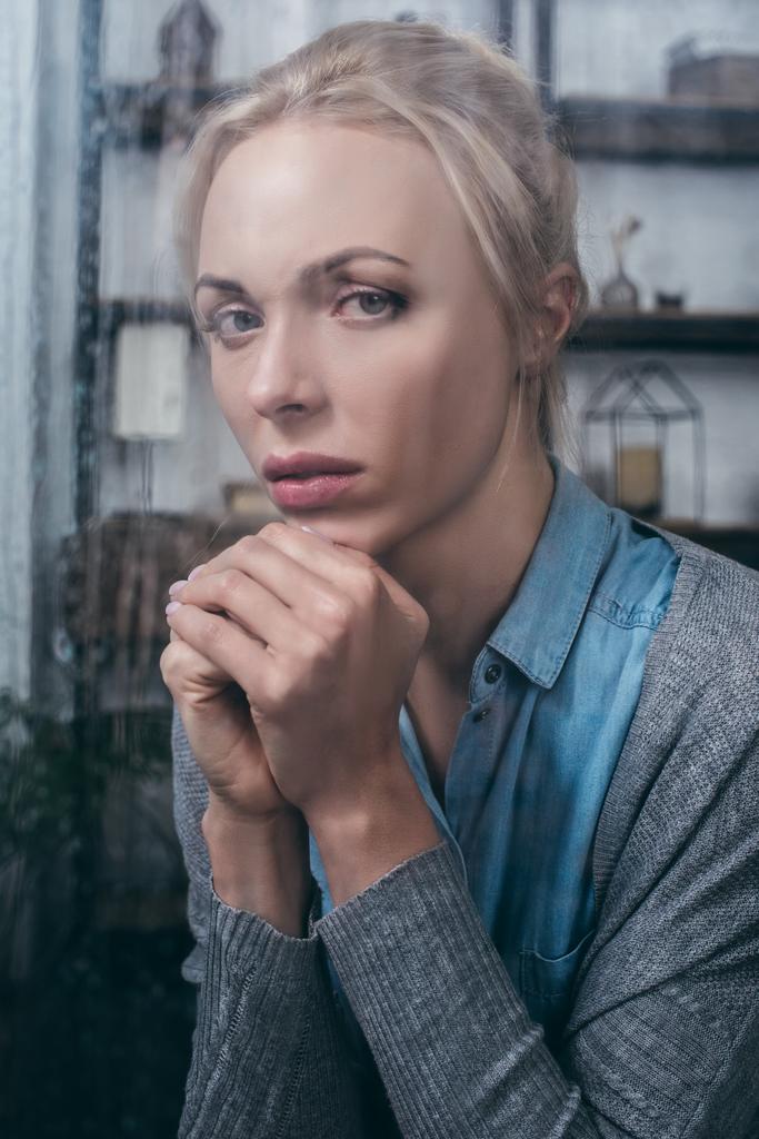 расстроенная взрослая женщина со сложенными руками, смотрящая в камеру дома через окно с капельками дождя
 - Фото, изображение