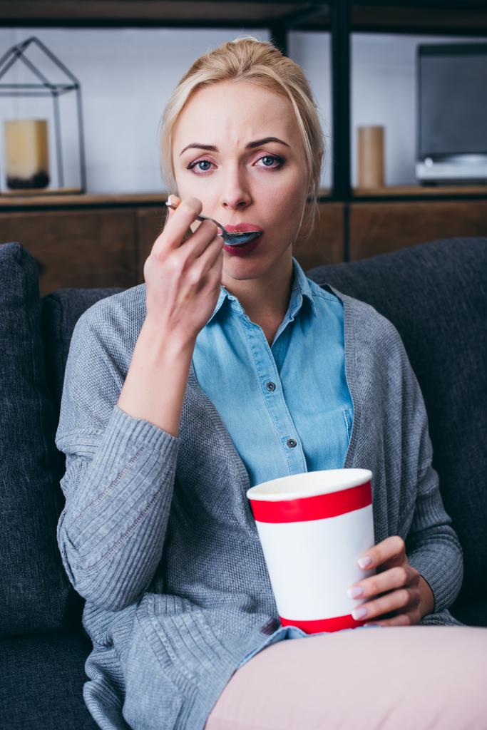Traurige Frau blickt in die Kamera und isst Eis, während sie allein zu Hause auf der Couch sitzt - Foto, Bild