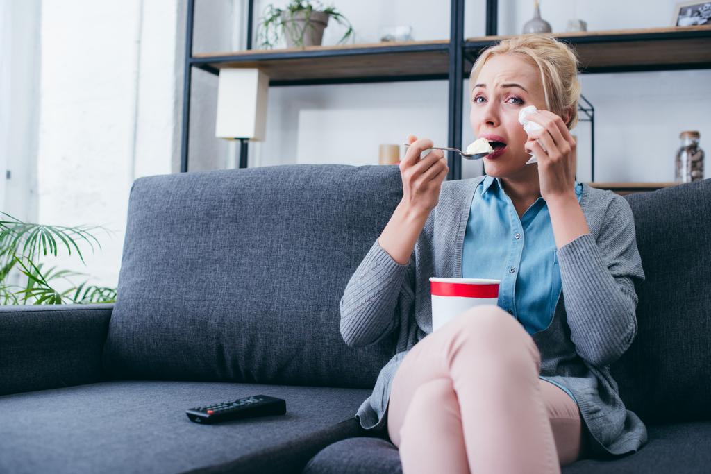 woman eating ice cream and crying while siitng on couch and watching tv at home alone - Photo, Image