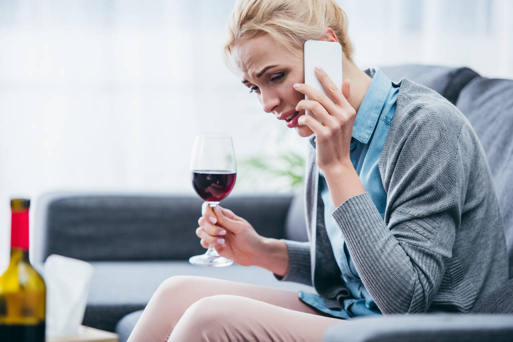 Verärgerte Frau sitzt auf Couch, hält Glas Rotwein in der Hand und telefoniert zu Hause mit Smartphone - Foto, Bild