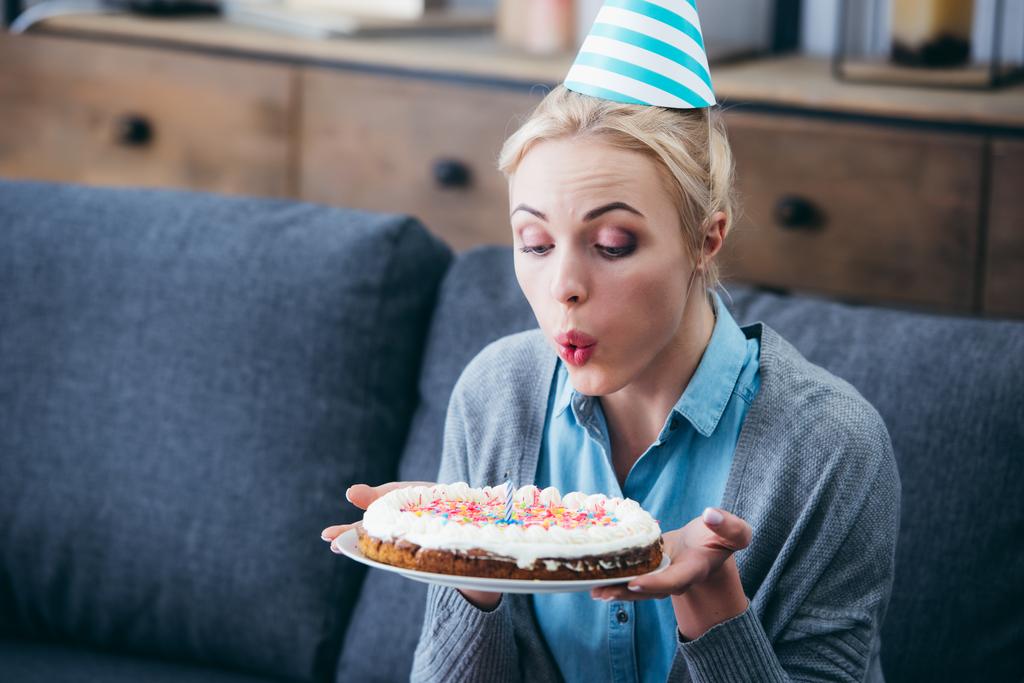γυναίκα με καπέλο κόμμα φυσώντας κερί για κέικ ενώ γιορτάζει τα γενέθλια στο σπίτι μόνος - Φωτογραφία, εικόνα