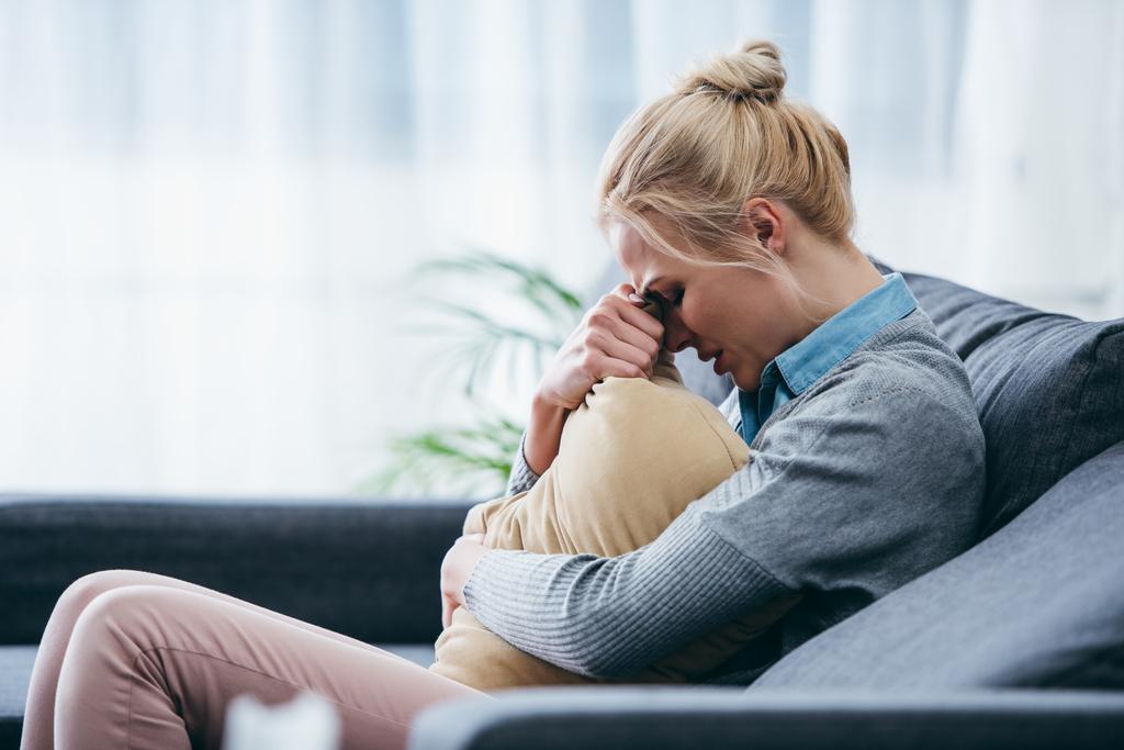 traurige Frau sitzt auf Couch, weint und hält Kissen zu Hause - Foto, Bild