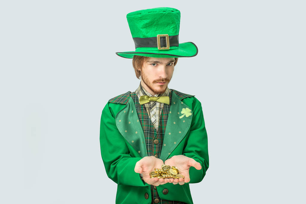 Νεαρός άνδρας στο πράσινο κοστούμι Κρατήστε χρυσά νομίσματα στα χέρια. Κοίταξε σοβαρά στη φωτογραφική μηχανή. Τύπος φορούν Αγίου Patricks κοστούμι. Απομονωμένα σε γκρι φόντο. - Φωτογραφία, εικόνα