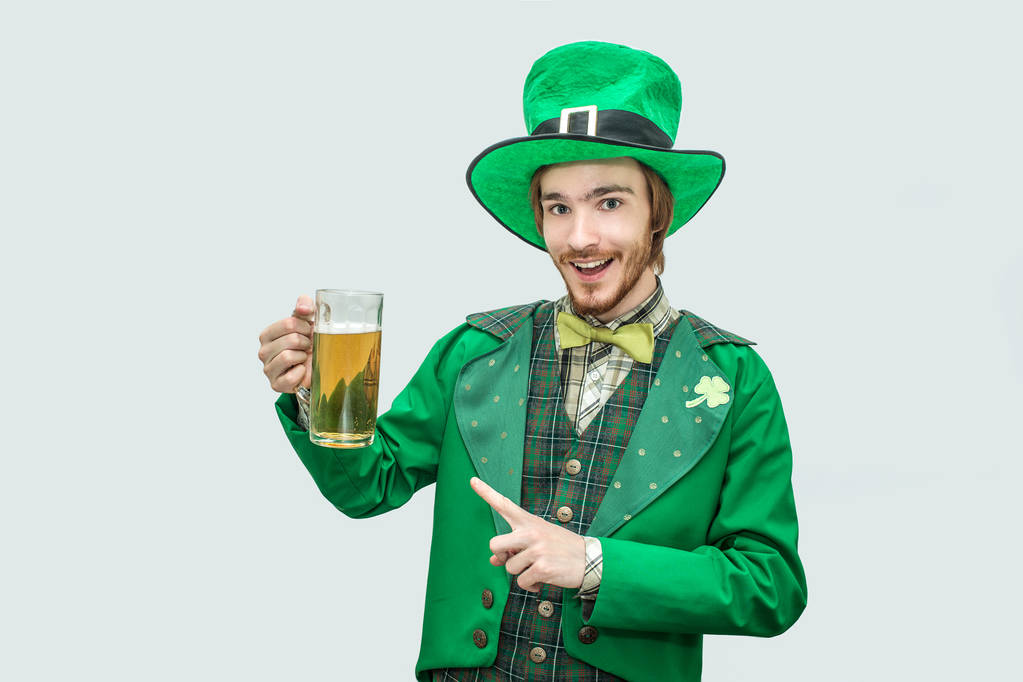 Onnellinen iloinen nuori mies vihreässä pyhimys patick puku tilalla muki olutta ja katso kameraan. Hän osoittaa sillä ja hymyilee. Eristetty harmaalla pohjalla
. - Valokuva, kuva