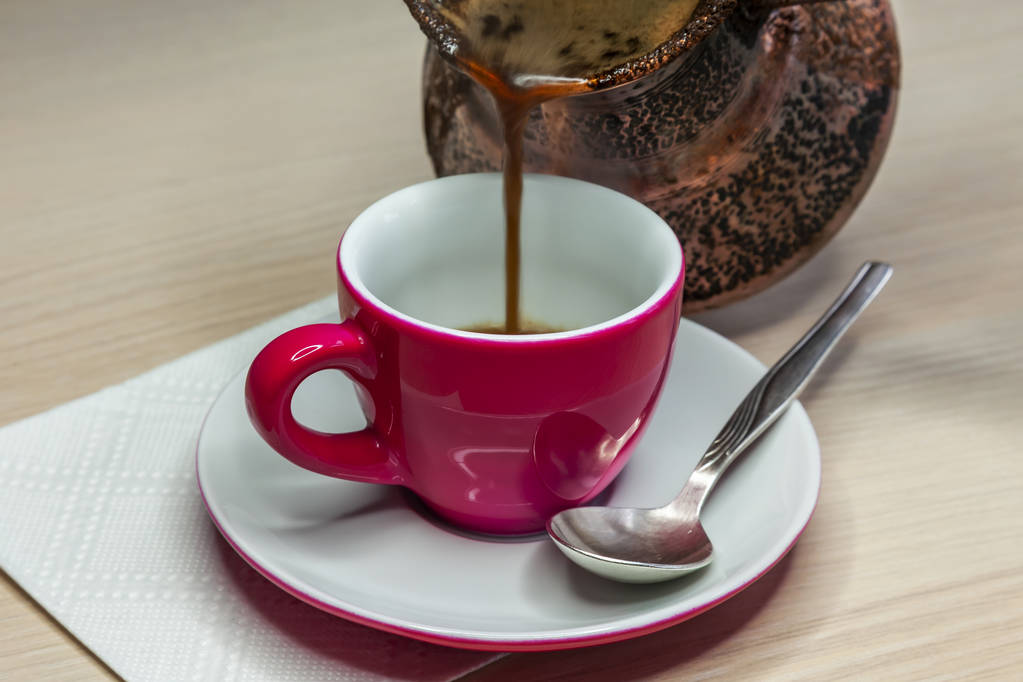 Zwarte koffie vloeit voort uit een cezve in een helder roze koffiekopje - Foto, afbeelding
