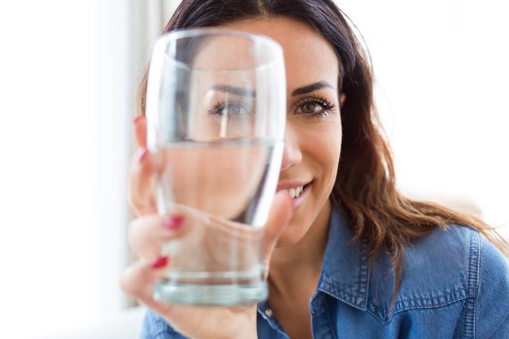Retrato de uma jovem mulher bonita sorrindo enquanto olha para a câmera através do copo de água em casa
. - Foto, Imagem