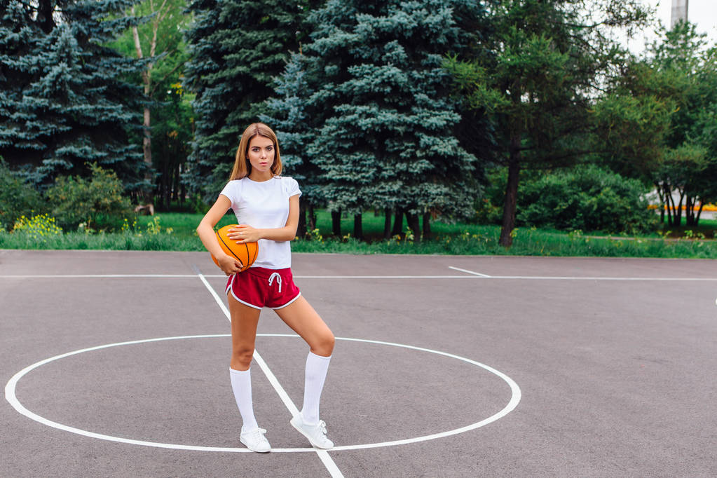 白いTシャツ、ショートパンツ、スニーカーを着た美しい若いブロンドの女の子は、屋外のバスケットボールコートでボールで遊んでいます。スペースのコピー. - 写真・画像