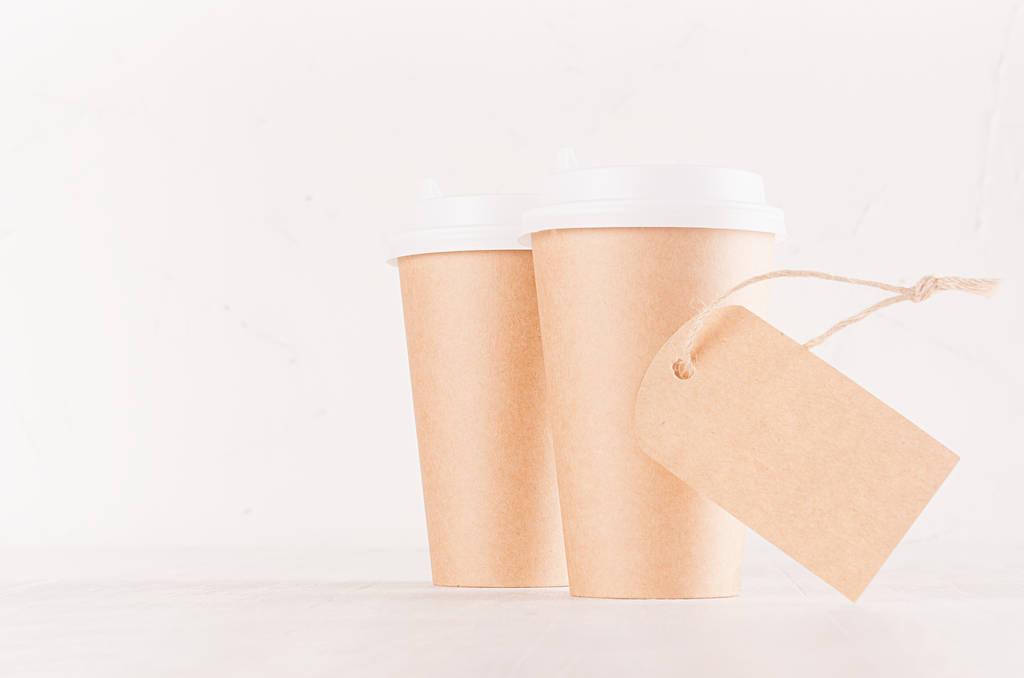 Kávé csomagoló makett - két kézműves barna papír cups fehér sapka és üres label Közelkép a fehér fa fedélzet-val másol hely, belső kávézóban. Modern elegáns koncepció a márka identitását, reklám, design. - Fotó, kép
