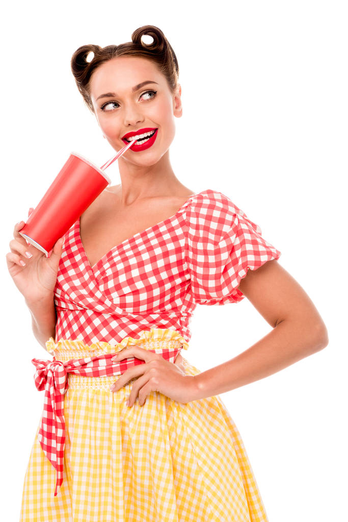 Smiling pin up girl drinking from paper cup with straw and keeping hand on hip isolated on white - Photo, Image