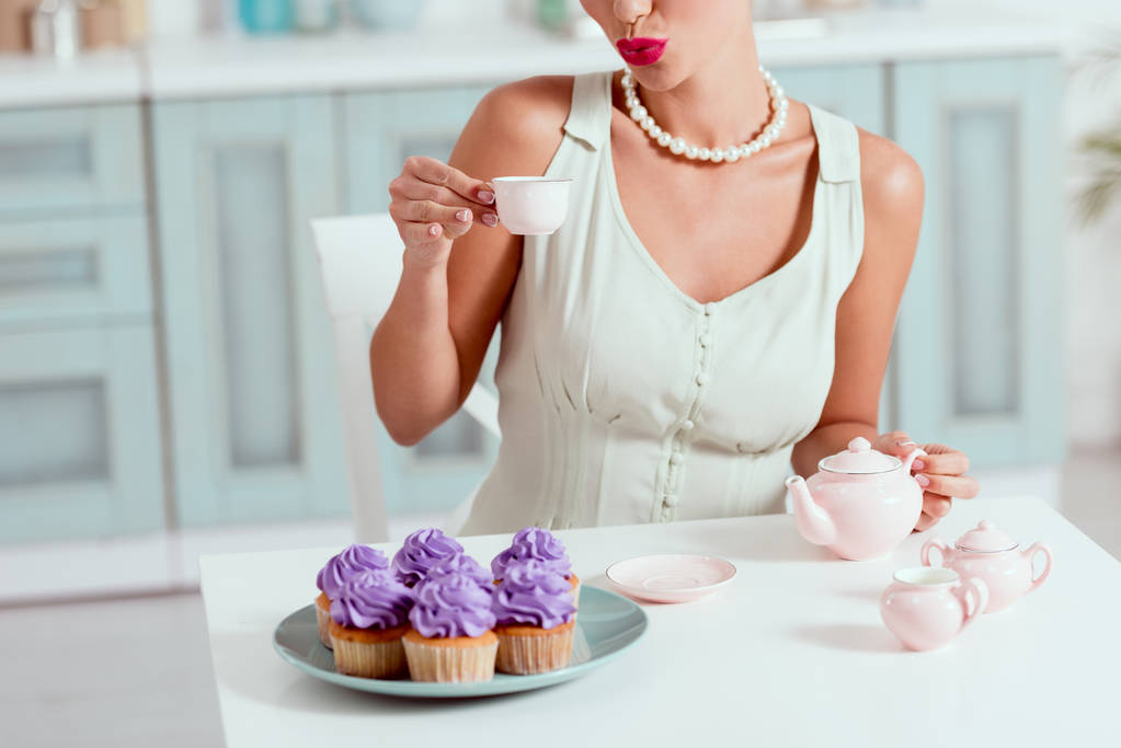 abgeschnittene Ansicht des Pin-up-Mädchens mit einer Tasse Kaffee am Tisch sitzend mit einem Teller Cupcakes - Foto, Bild