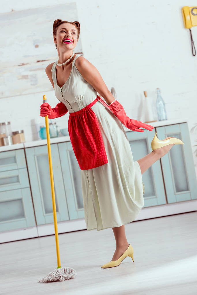 Smiling pin up girl in red apron and red rubber gloves dancing with yellow mop - Photo, Image