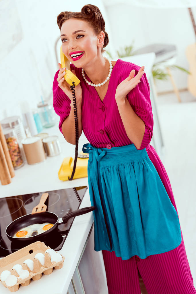 Pin-up lány, sütés, tojás, és beszélni, vintage sárga telefon mosolyogva - Fotó, kép