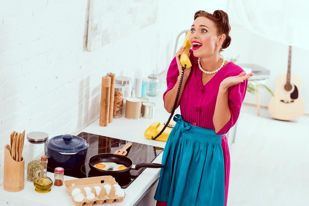 Excited pin up girl talkin on vintage yellow phone while frying eggs - Photo, Image