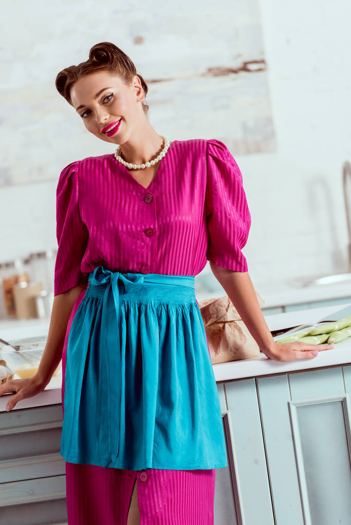 Elegante pin up girl en vestido carmesí y delantal azul de pie junto a la mesa de la cocina
 - Foto, imagen
