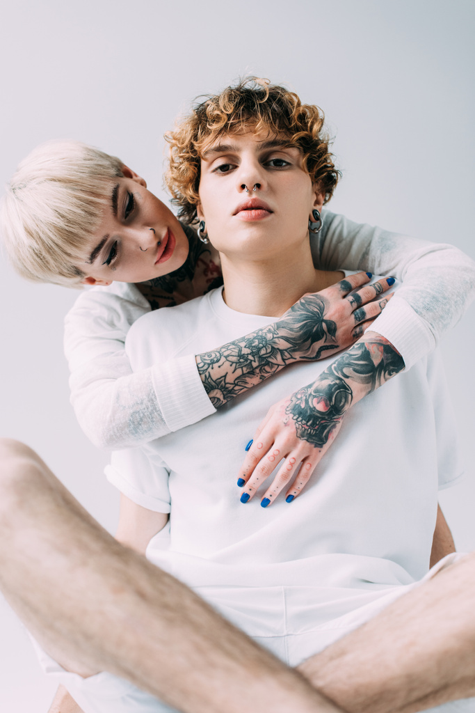 ξανθό κορίτσι με τατουάζ που αγκαλιάζει το φίλο με σγουρά μαλλιά που απομονώνονται σε γκρι - Φωτογραφία, εικόνα