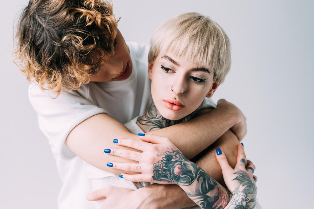 man met krullend haar omhelst blonde vrouw met tatoeages geïsoleerd op grijs - Foto, afbeelding