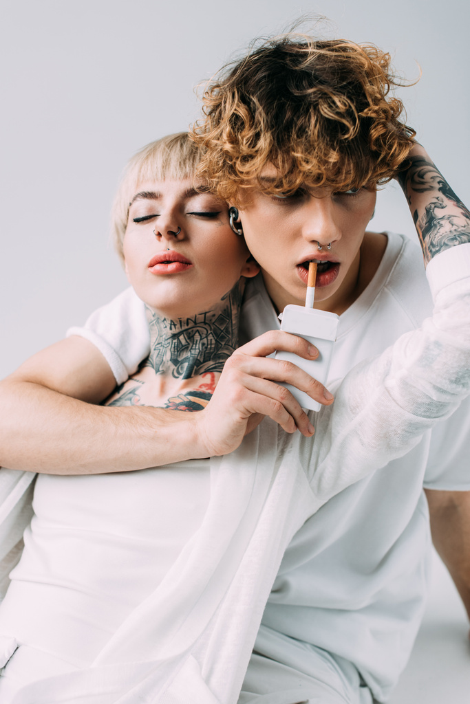 mujer tatuada abrazando hombre guapo con pelo rizado sosteniendo cigarrillo en la boca aislado en gris
  - Foto, imagen