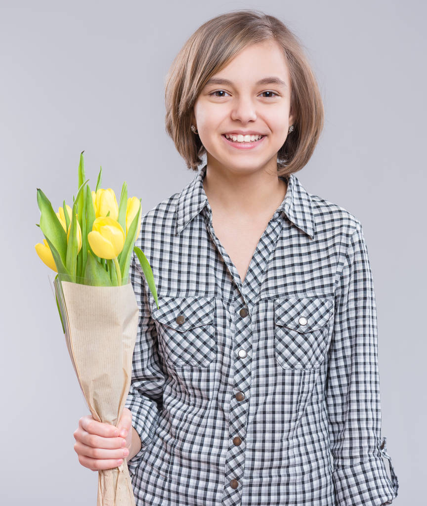 schönes Mädchen mit einem Strauß Tulpen auf grauem Hintergrund. Teenie-Mädchen hat eine Überraschung für Mama vorbereitet - gelbe Blumen. Lustiger Teenager, der in die Kamera lächelt. - Foto, Bild
