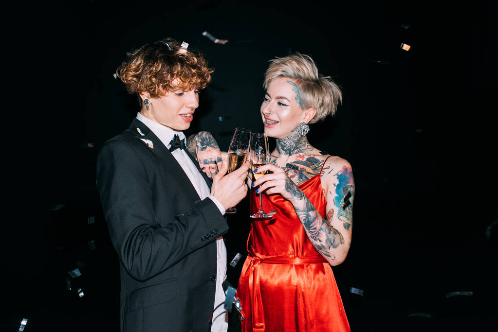 vrolijke man met krullend haar roosteren met glas champagne met gelukkige vrouw geïsoleerd op zwart  - Foto, afbeelding