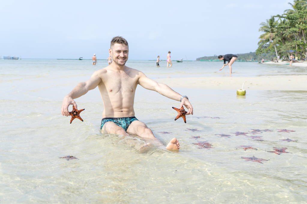 Cestovní chlapec sedí na moře s červenými starfishes dělá legrační fotografie v Ostrově Phu Quoc v křišťálově čisté vodě - Fotografie, Obrázek
