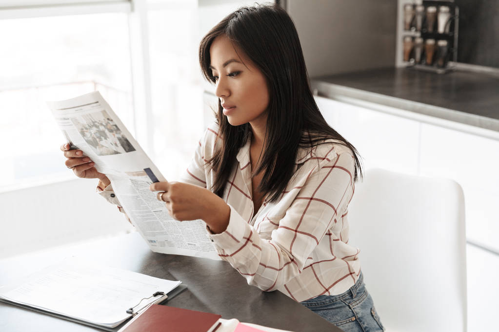 Fiatal ázsiai nő 20s olvasó újság fedett asztalnál ülve képe - Fotó, kép
