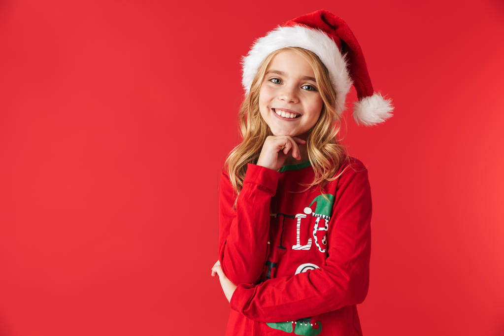 Веселая маленькая девочка в рождественском костюме стояла изолированная на красном фоне, смотрела в камеру
 - Фото, изображение
