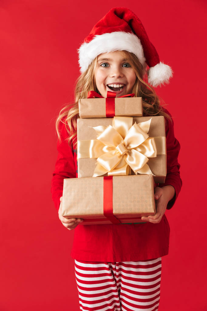Веселая маленькая девочка в рождественском костюме стояла изолированная на красном фоне, держа подарочные коробки
 - Фото, изображение