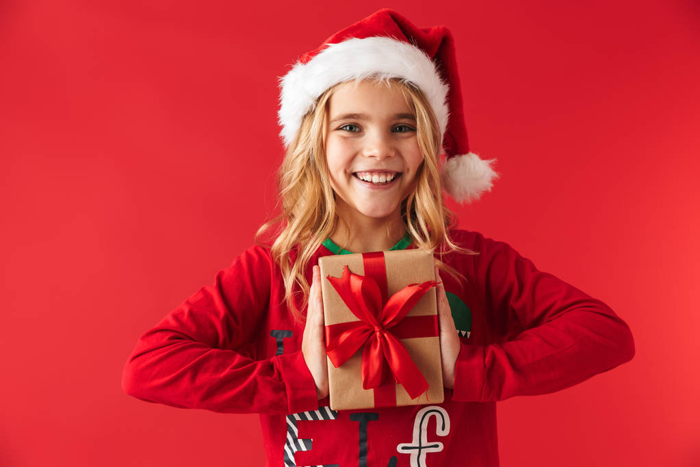 Noel şapka ayakta giyen sevimli küçük kız kırmızı arka plan üzerinde mevcut kutusu tutarak izole - Fotoğraf, Görsel