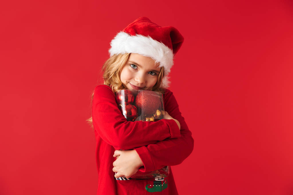 Cheerful little girl wearing Christmas costume standing isolated over red background, holding set of Christmas tree toys - Photo, Image
