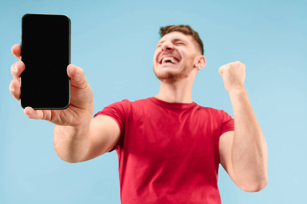 Joven hombre guapo mostrando la pantalla del teléfono inteligente aislado en el fondo azul en shock con una cara sorpresa
 - Foto, Imagen