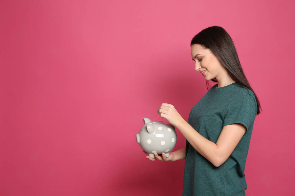 Νεαρή γυναίκα βάζοντας κέρμα σε τράπεζα piggy σε έγχρωμο φόντο. Χώρο για το κείμενο - Φωτογραφία, εικόνα