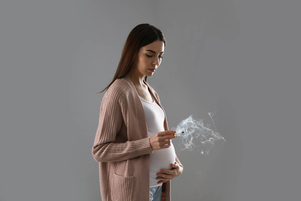 Young pregnant woman smoking cigarette on grey background - Photo, Image