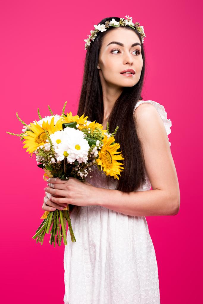 mooie brunette vrouw in witte jurk en bloemen krans holding bloemboeket en op zoek weg geïsoleerd op roze - Foto, afbeelding