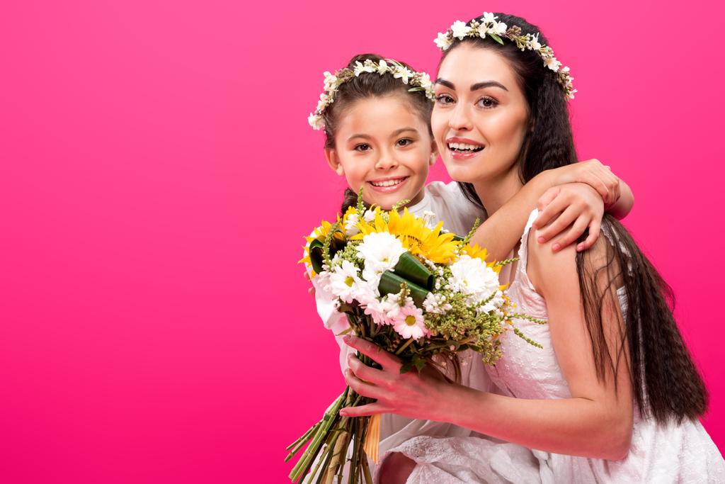 beautiful happy mother and daughter with flower bouquet hugging and smiling at camera isolated on pink - Photo, Image