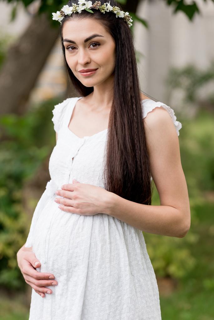beautiful happy young pregnant woman in white dress and floral wreath smiling at camera in park - Photo, Image